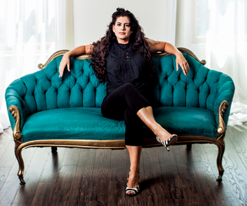 Katie Knipp sitting on couch with arms spread and legs crossed, long dark curly wavy hair