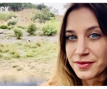 Trina Hill photo of face, blue eyes, straight brown hair, grassy field in background with a sign in background that spells, "luck"