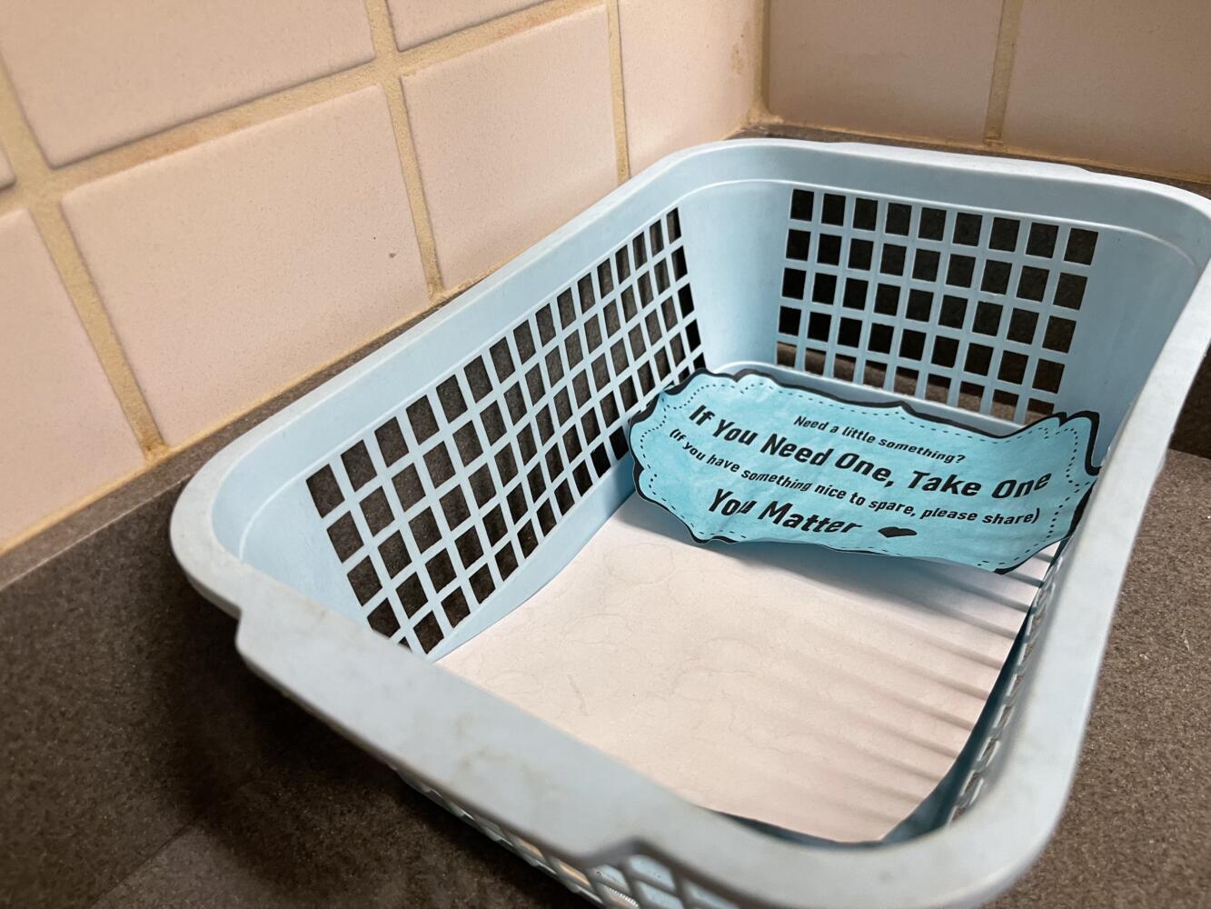 empty laundry basket on bathroom floor with paper sign in it, 'if you need one, take one, if you have one, give one'