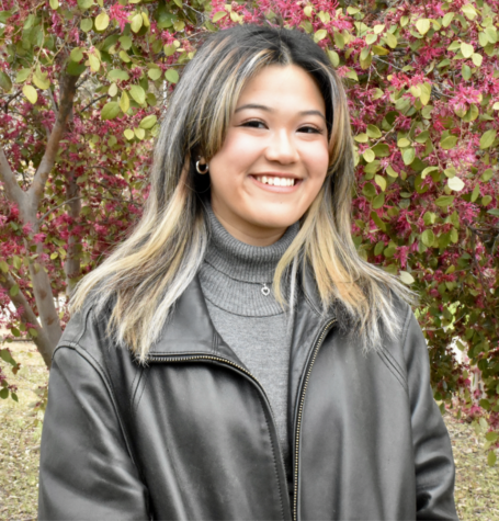 Photo of Alexa Topacio in a grey sweater and black leather coat, she is smiling and has long hair and a blooming tree is behind her.