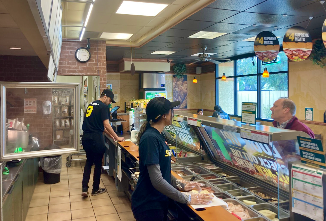 Subway store workers stand behind counter and take order to make sandwiches