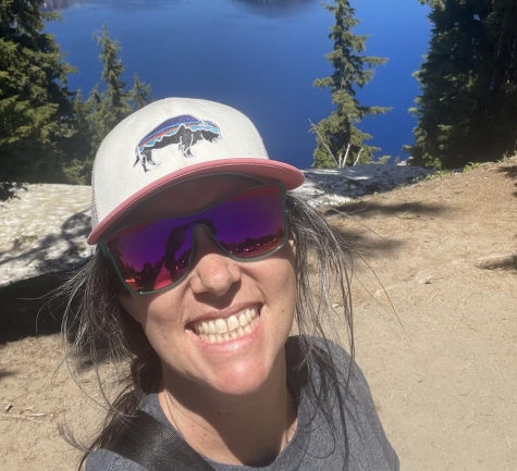 Rachel is smiling with a baseball hat and sunglasses on, standing in front of a lake